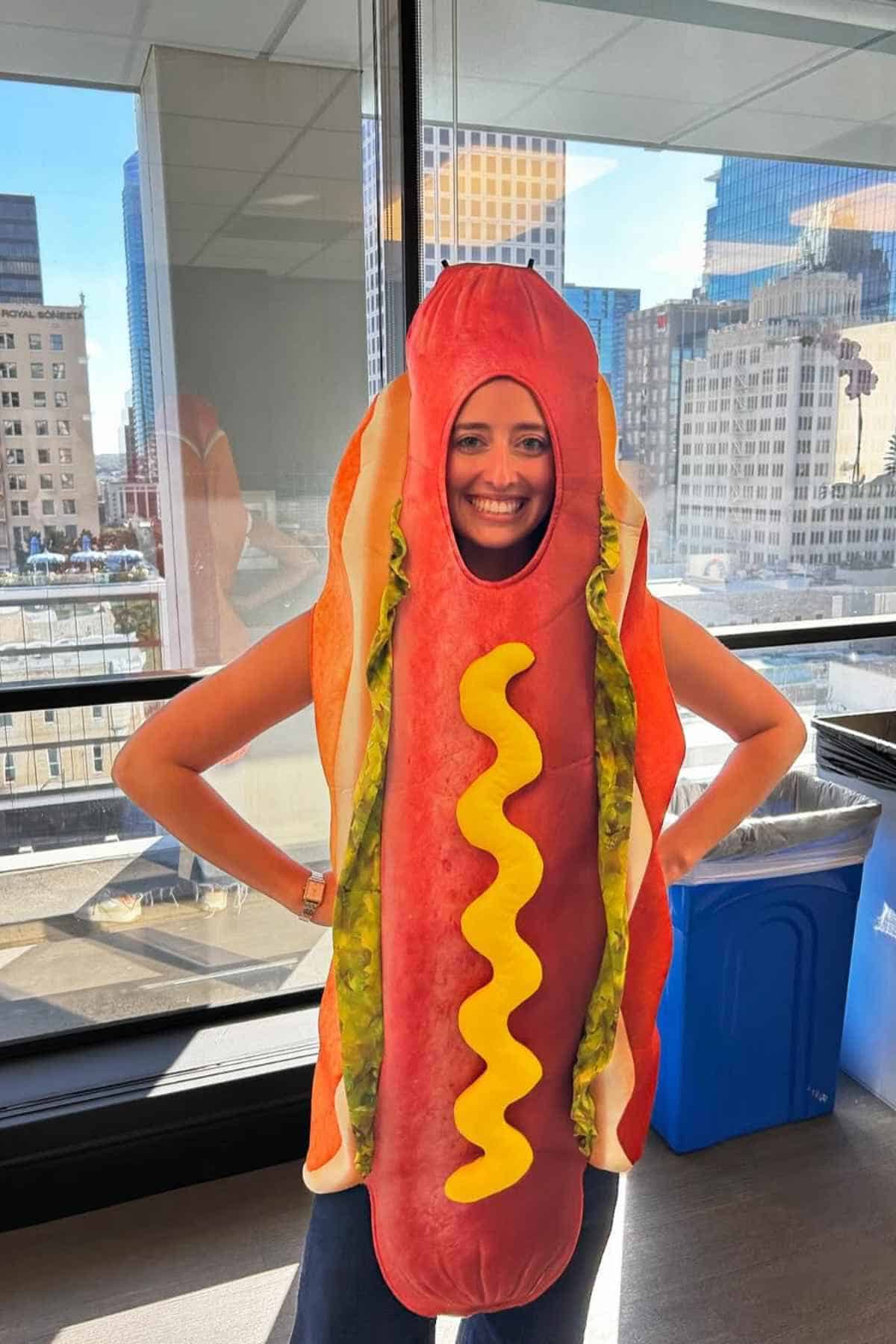 A person stands indoors wearing a hot dog costume with mustard, in front of large windows showing city buildings outside.
