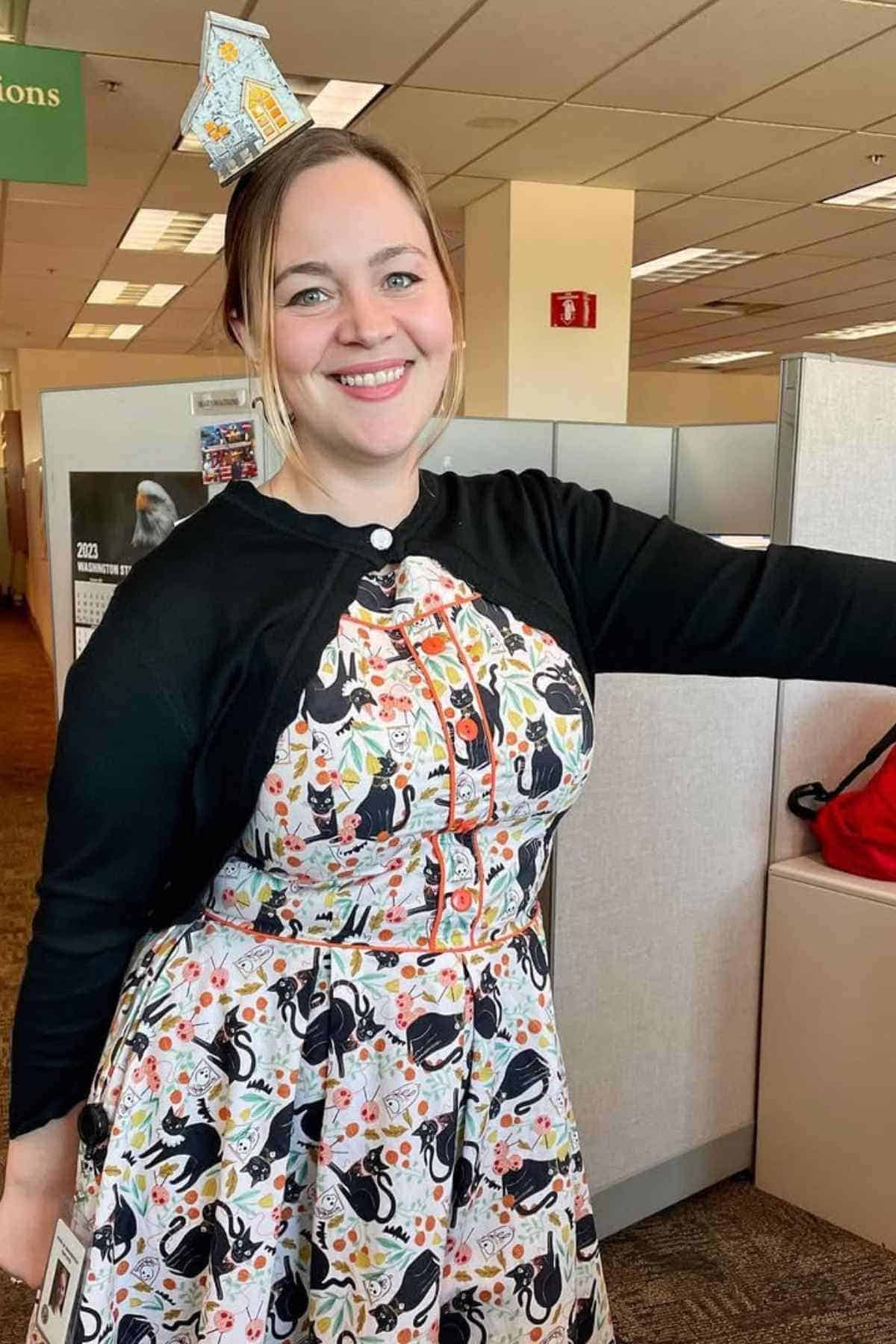 Woman standing in an office cubicle, smiling, wearing a colorful dress with a black cardigan and a paper house cat on her head.
