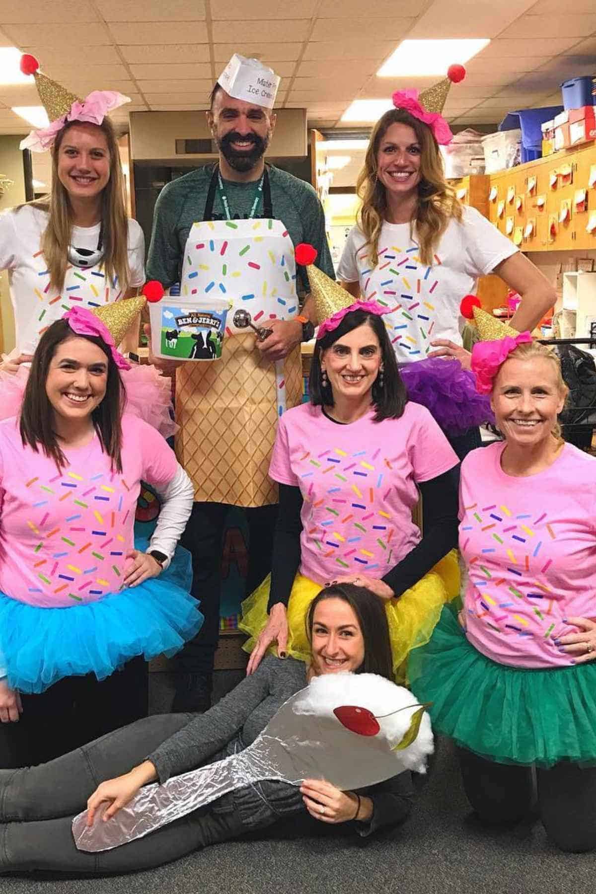 A group of seven adults dressed in ice cream and dessert-themed costumes pose and smile together in a brightly lit room.