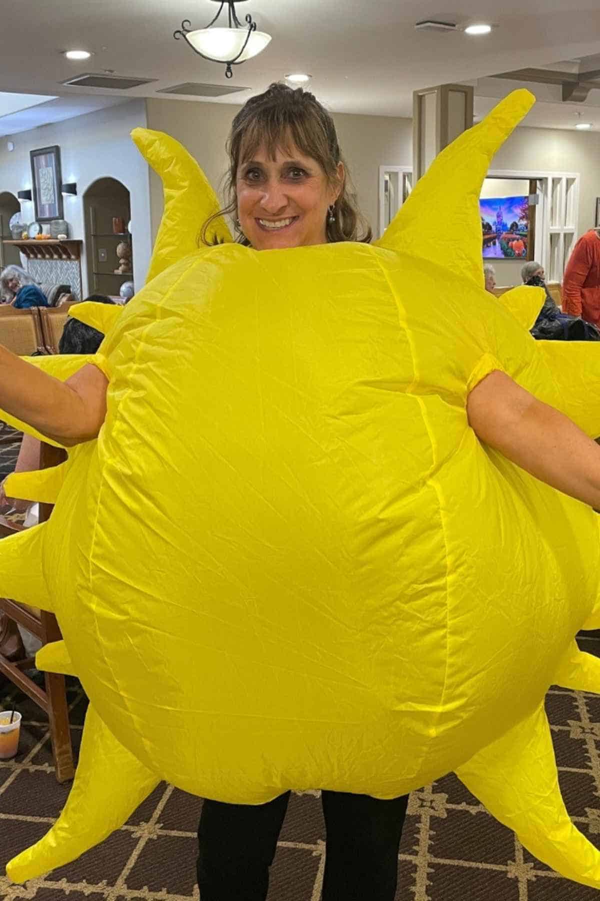 A woman smiles while wearing a large, inflatable yellow sun costume inside a room with other people in the background.