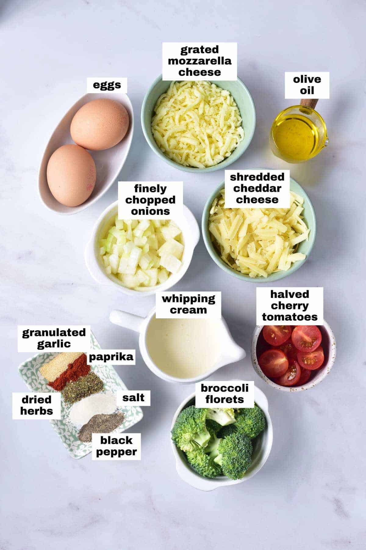 Assorted labeled ingredients on a white surface, including eggs, grated cheeses, olive oil, onions, whipping cream, cherry tomatoes, broccoli, garlic, spices, salt, and pepper.
