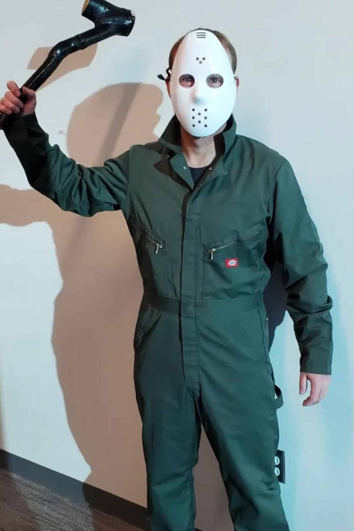 Person wearing a green jumpsuit and a white hockey mask, holding a black axe prop, standing against a plain wall.