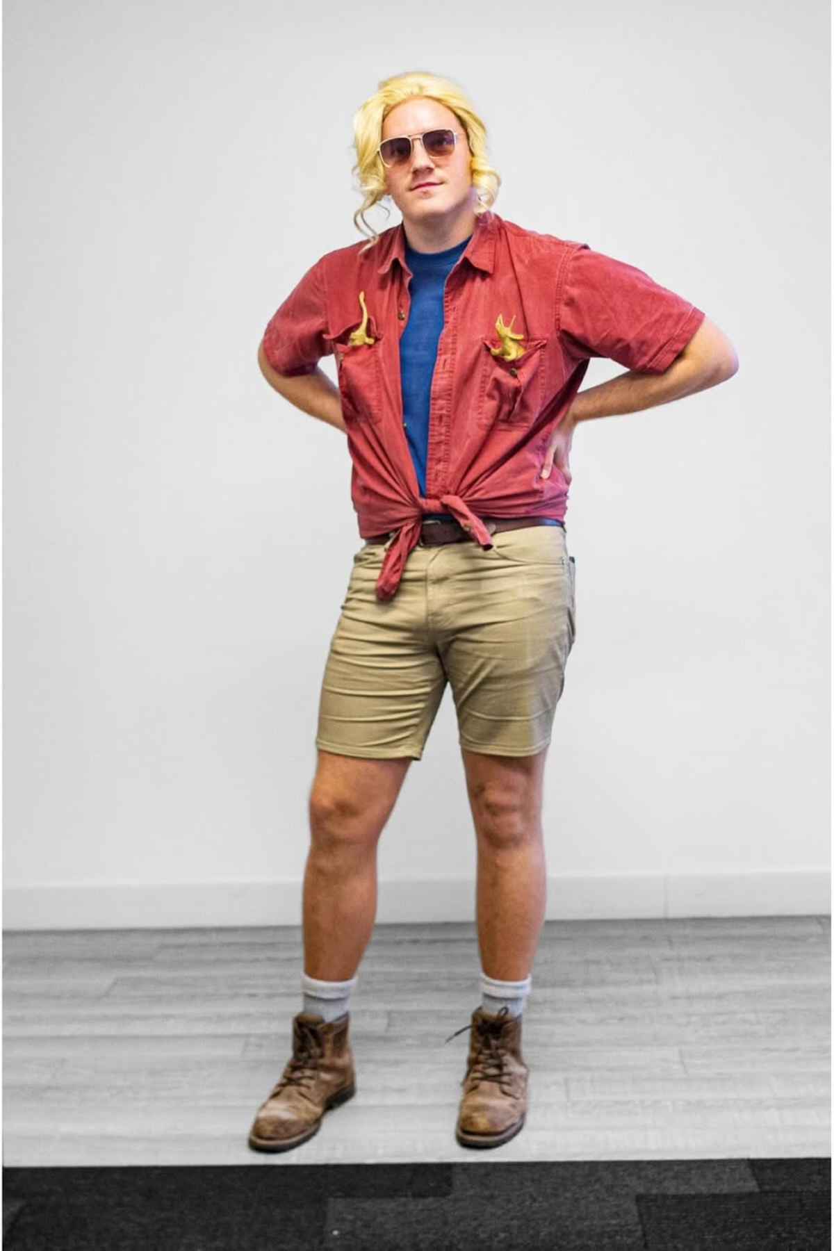 A person stands with hands on hips, wearing a blonde wig, aviator sunglasses, a red tied shirt with embroidered birds, a blue undershirt, khaki shorts, socks, and brown boots.