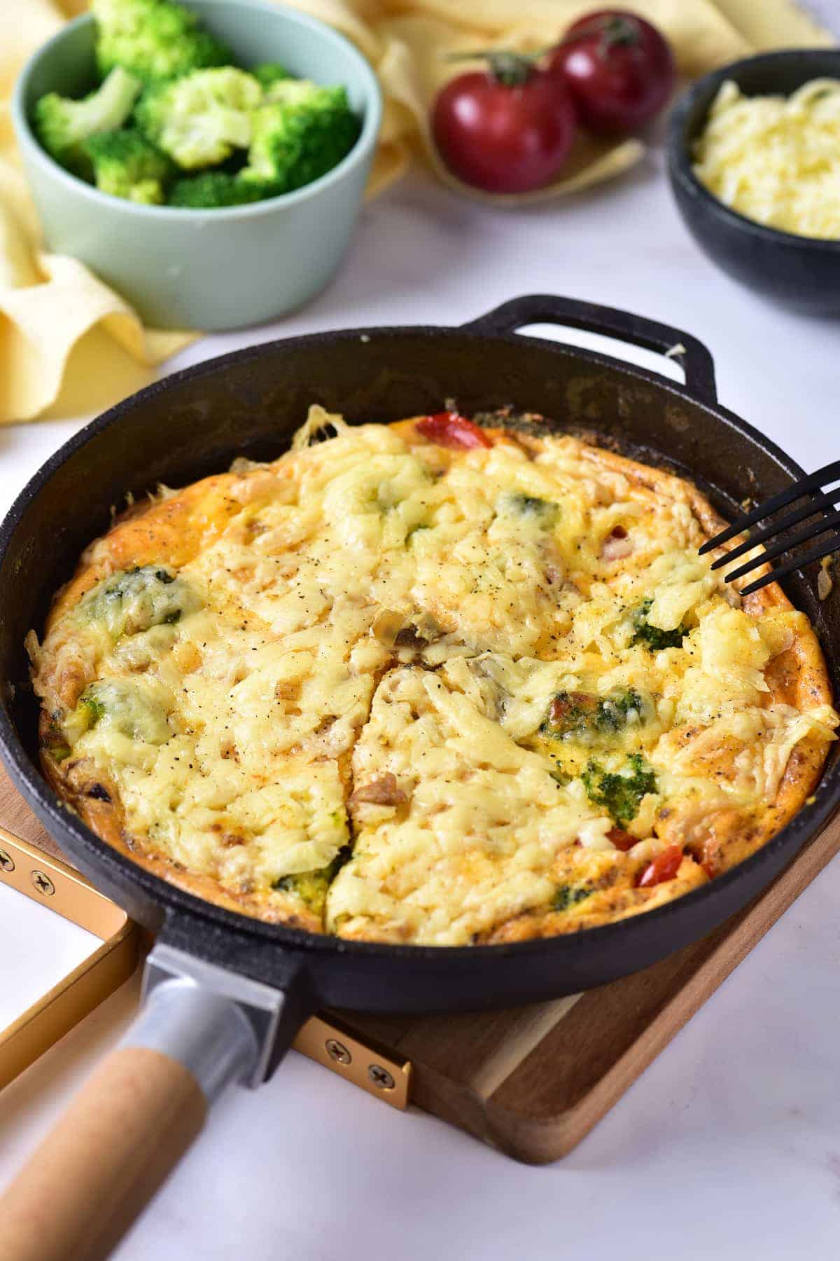 A baked vegetable frittata with melted cheese in a cast iron skillet, with broccoli, tomatoes, and cheese in bowls nearby.