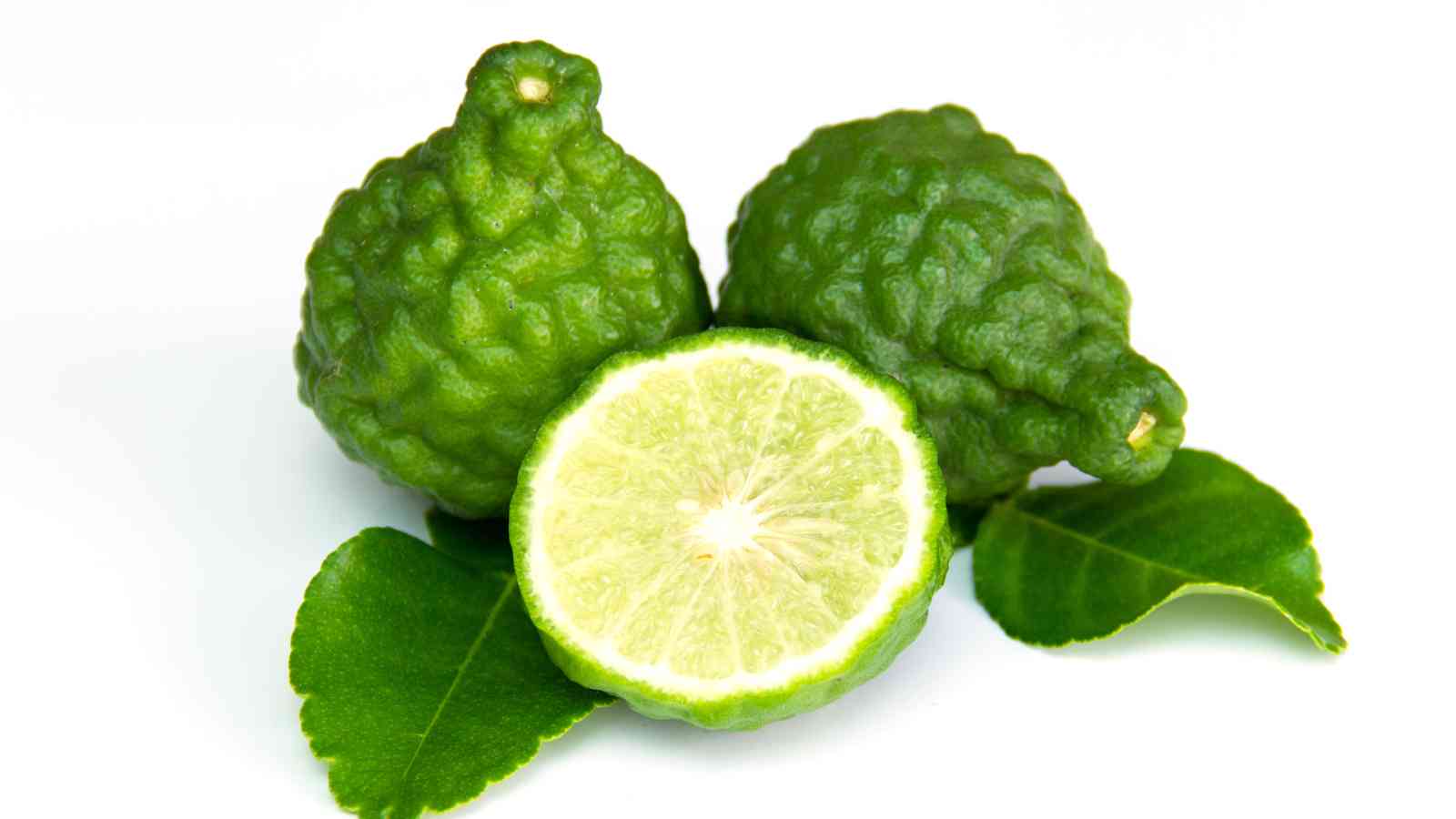 Three kaffir limes, two whole and one cut in half, rest on green leaves against a white background.