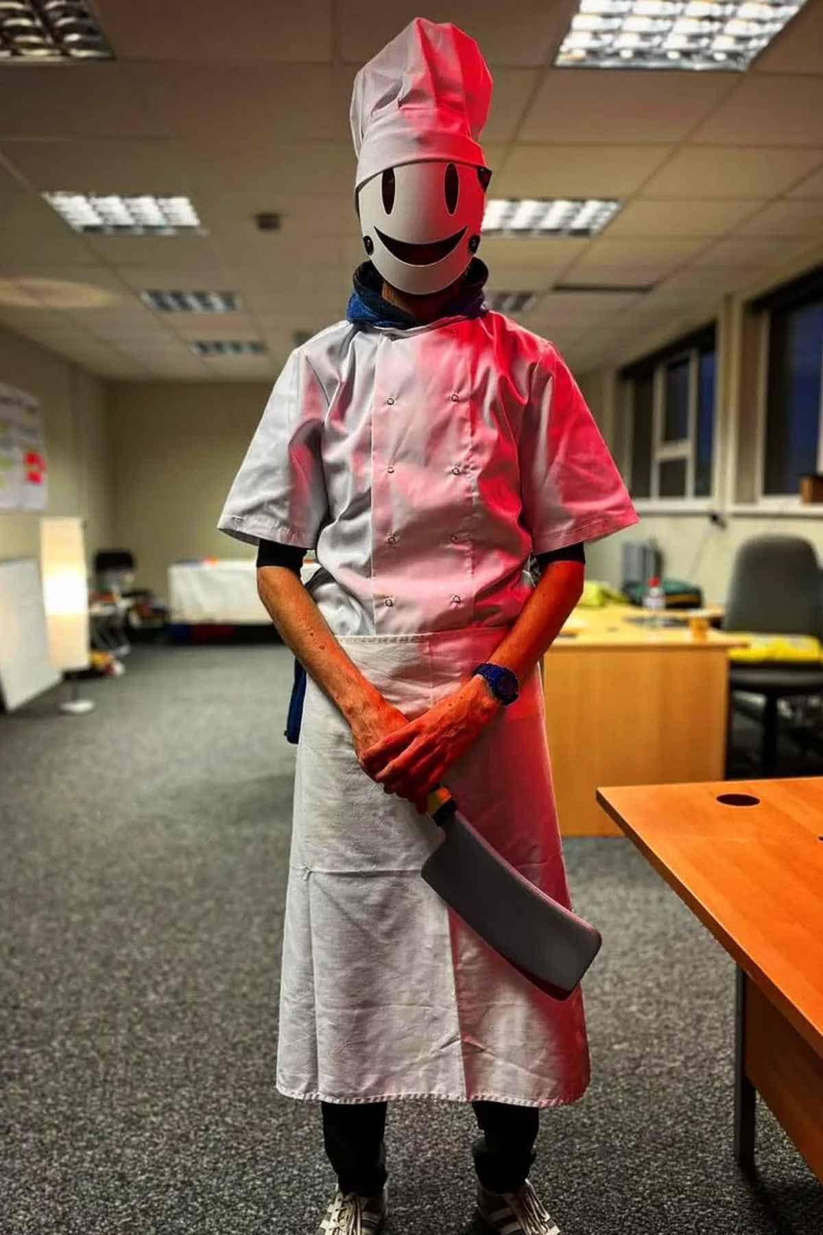 A person wearing a white chef outfit, tall hat, and a smiley face mask stands indoors holding a large cleaver in both hands.