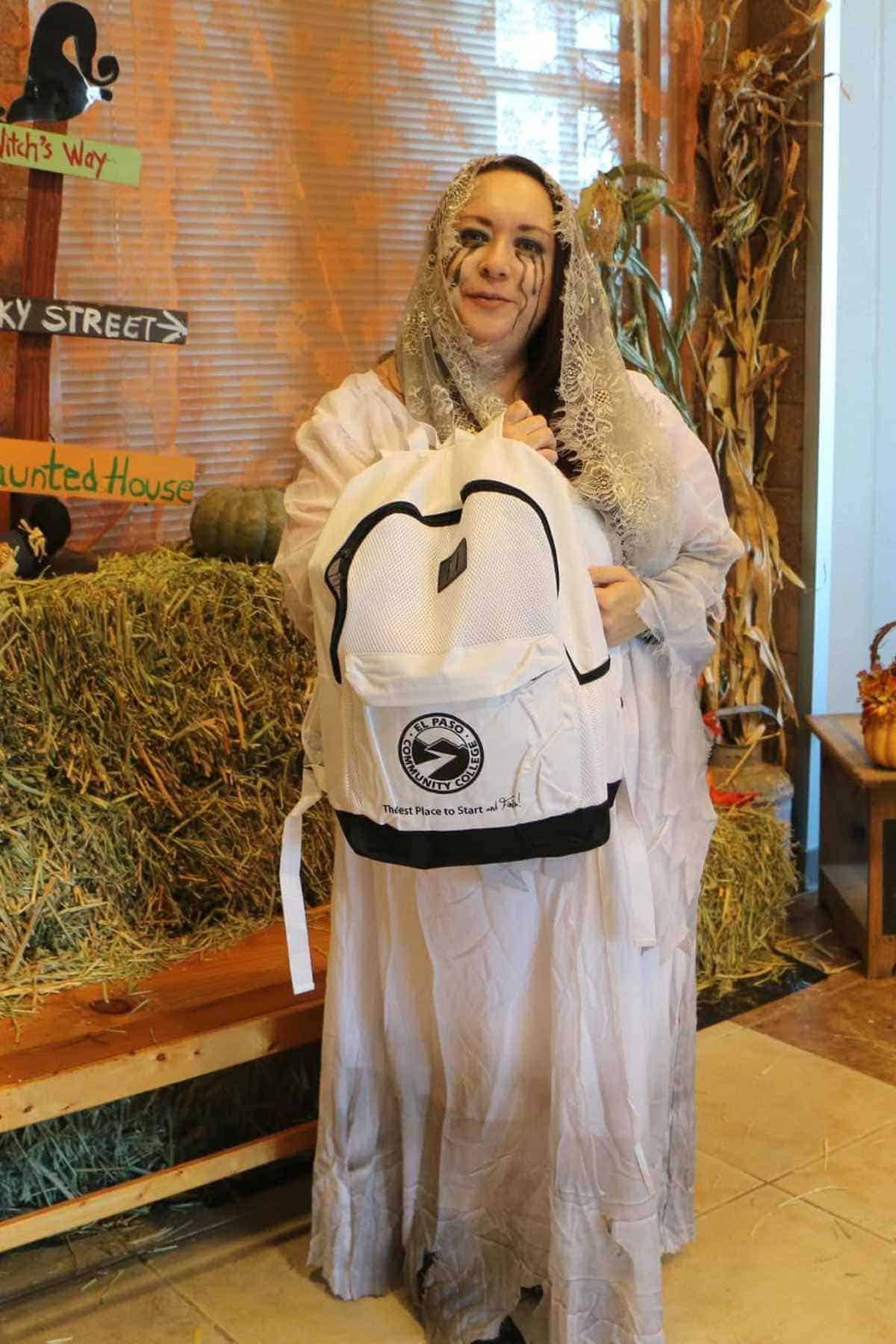 A person in a white ghost costume stands indoors holding a white backpack, with Halloween decorations and hay bales in the background.