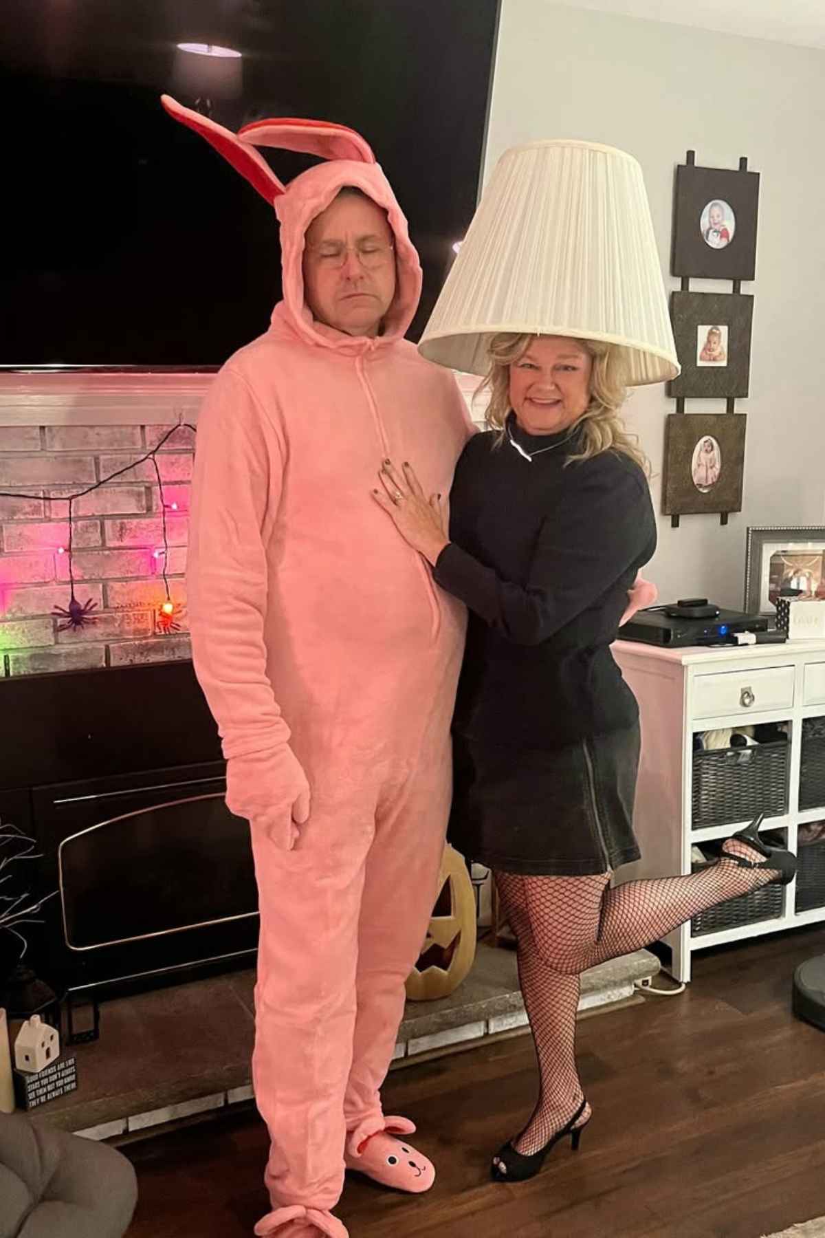 Two adults pose indoors; one is dressed in a pink bunny costume, and the other wears a lampshade on their head, standing with one foot raised and a hand on the other's chest.