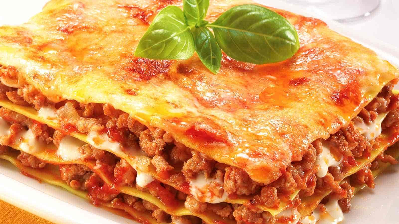 A close-up of a slice of baked lasagna with layers of pasta, ground meat, tomato sauce, and melted cheese, topped with a sprig of fresh basil.