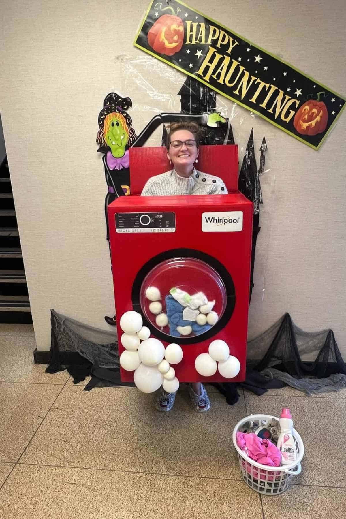 Person wearing a washing machine costume with soap bubbles stands in front of Halloween decorations, including a "Happy Haunting" sign and a laundry basket with clothes.