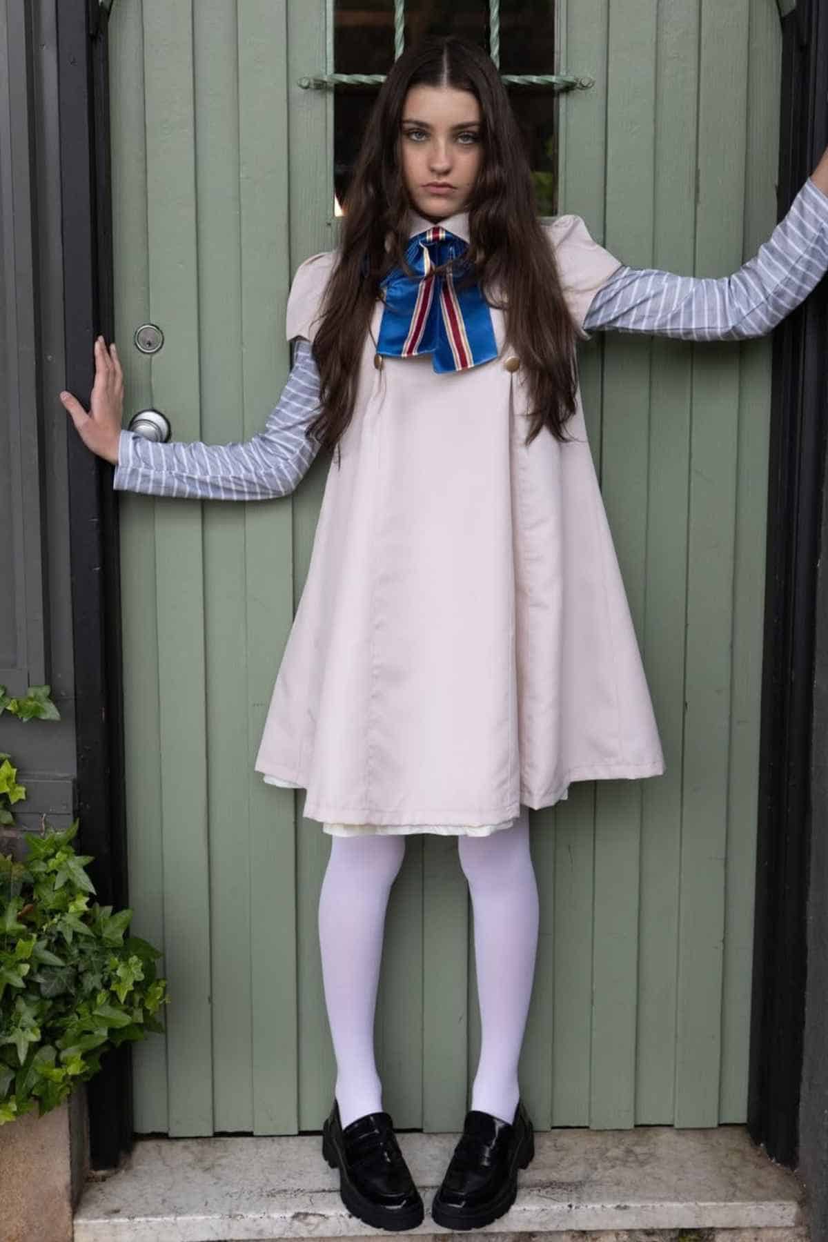 A woman stands in front of a green wooden door, wearing a white dress over a striped long-sleeve shirt, white tights, black shoes, and a blue ribbon with red and white stripes.