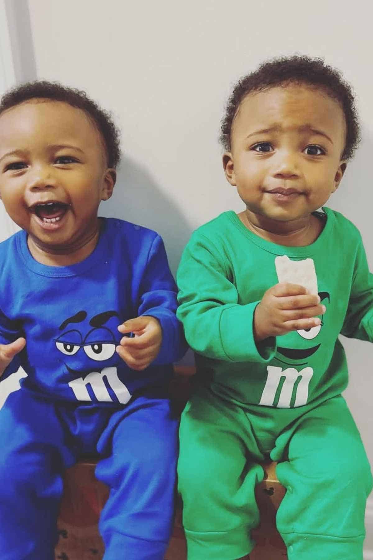 Two kids sit side by side wearing blue and green M&M pajamas. One child is laughing, while the other holds a white object and looks at the camera.