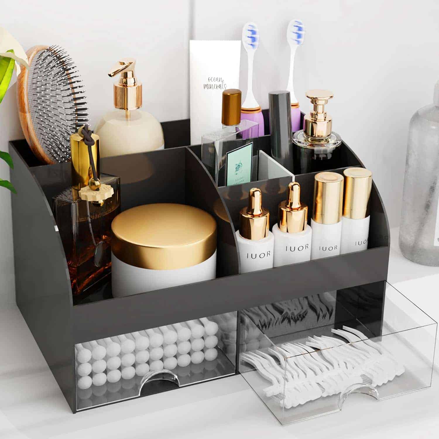A black tabletop organizer holding skincare products, lotions, a hairbrush, toothbrushes, cotton swabs, and floss picks, with two clear drawers partially open.