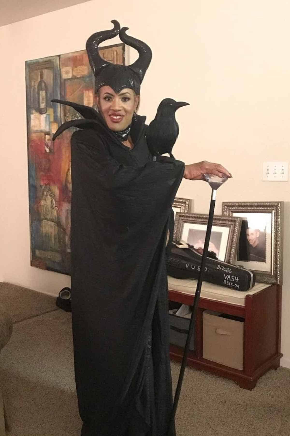 Person dressed as Maleficent in a black robe and horned headpiece, holding a staff with a black bird perched on their arm, standing in a living room.