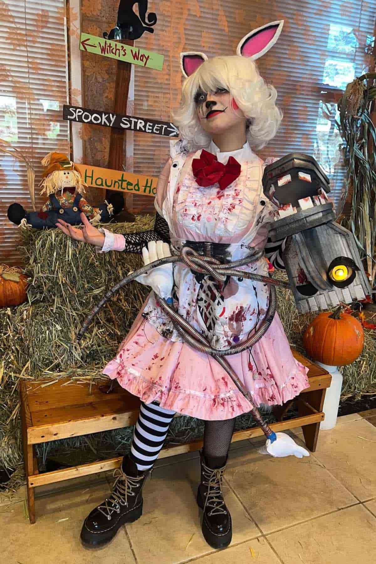 Person in a pink, blood-splattered costume with bunny ears, holding a doll and a prop weapon, stands in front of Halloween decorations with hay, pumpkins, and themed signs.