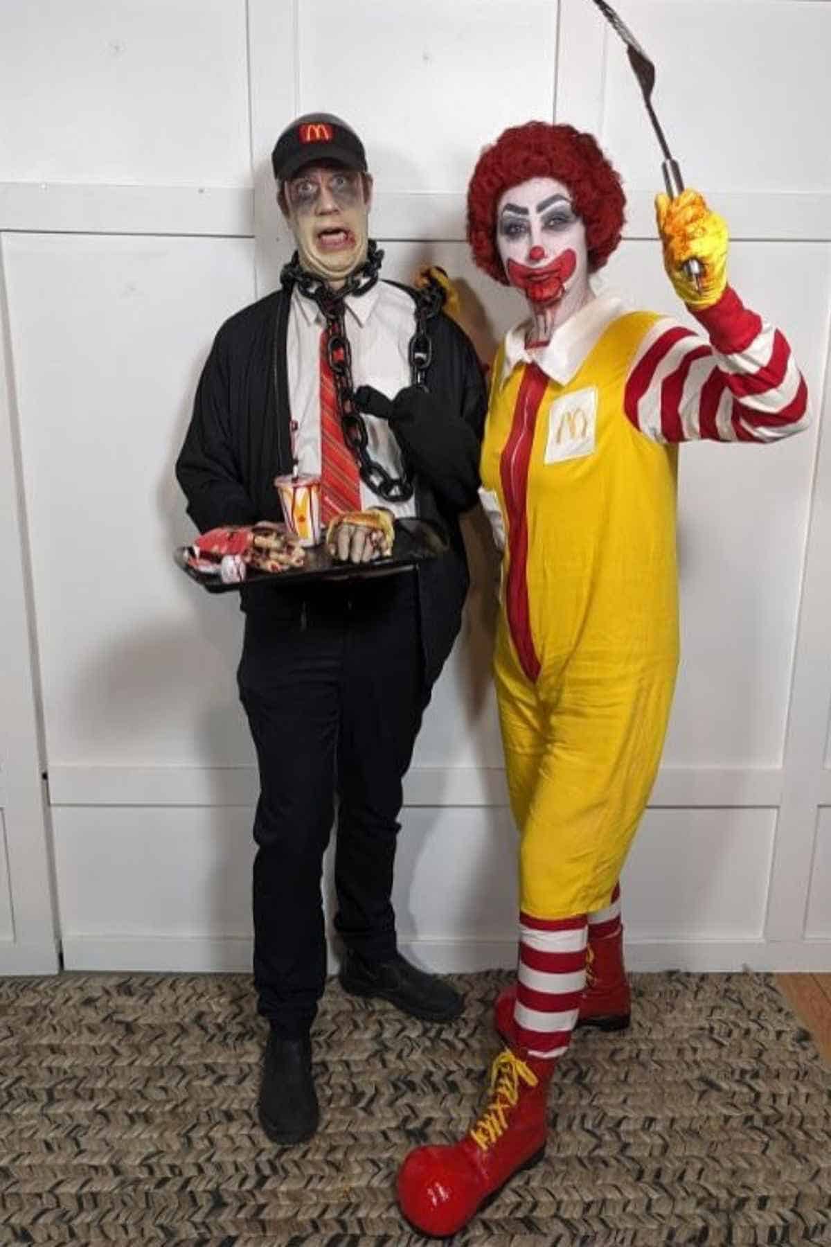 Two people dressed in McDonald's-themed costumes, one as a zombie employee holding food, and the other as Ronald McDonald holding a spatula, stand side by side indoors.