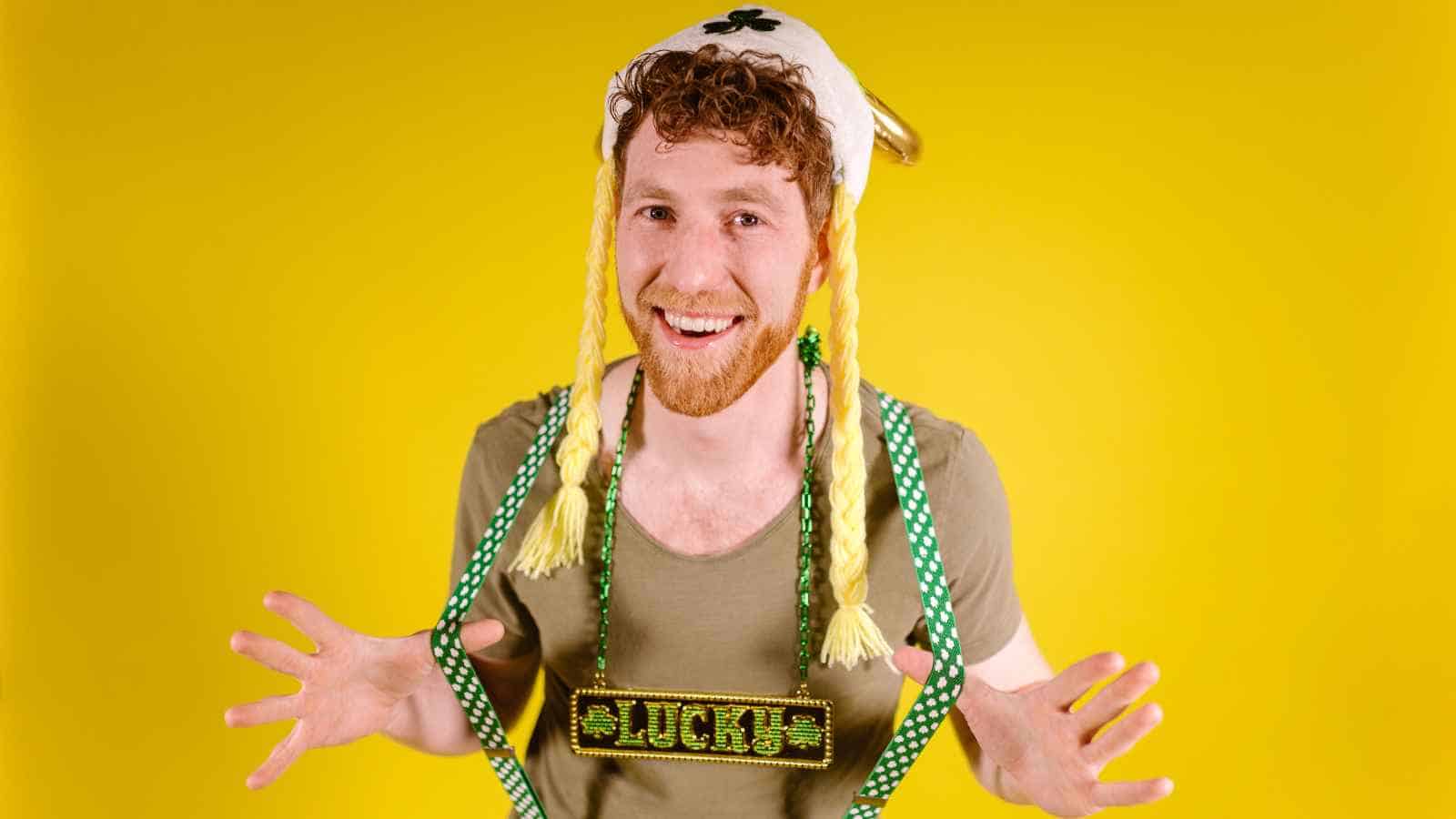 A smiling person with a fake blonde braided hat and a "lucky" necklace poses against a bright yellow background.