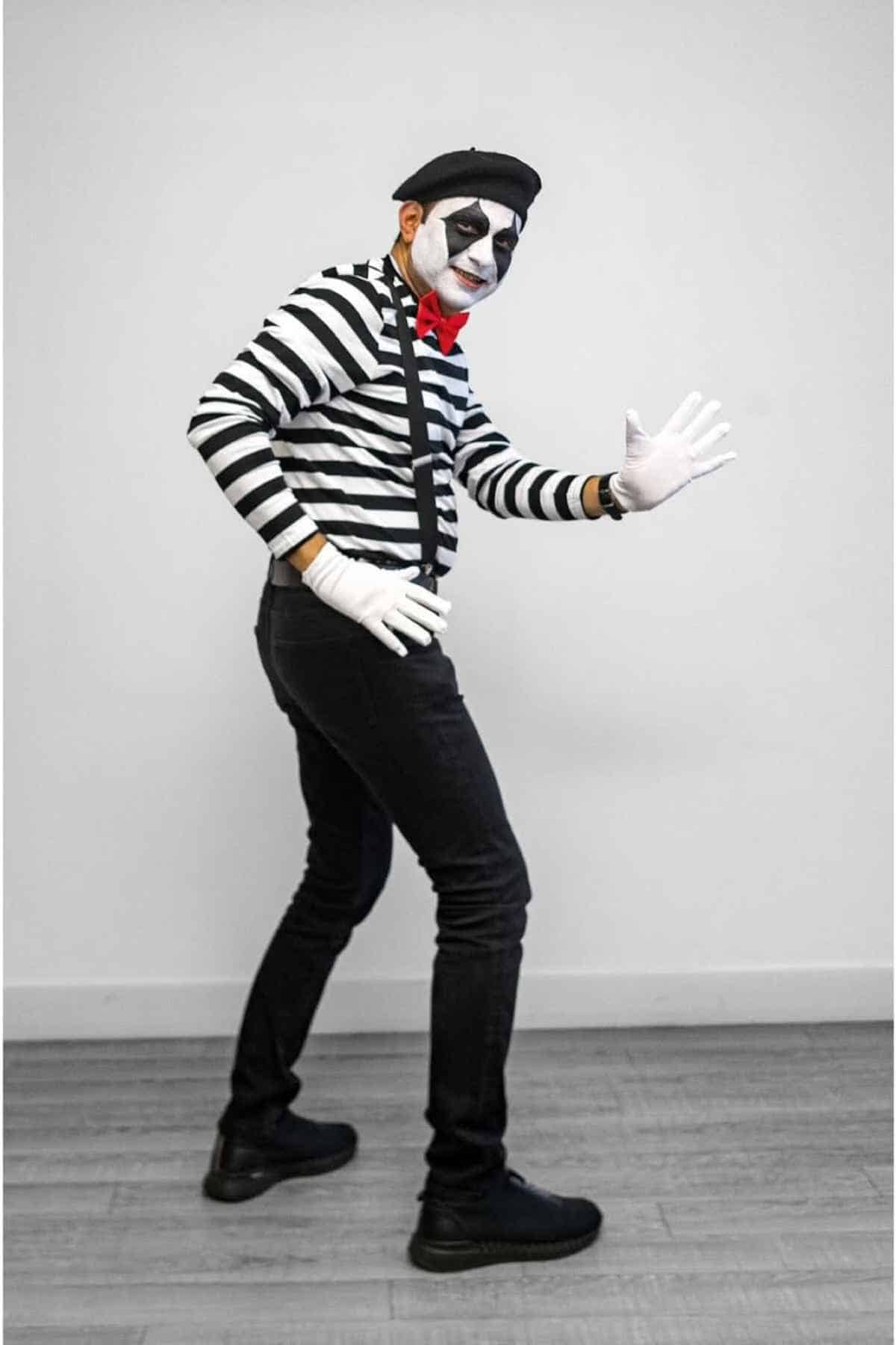Person dressed as a mime in a black beret, black and white striped shirt, white gloves, red bowtie, black pants, and black shoes, posing against a plain indoor background.