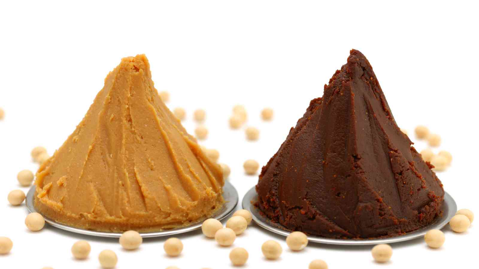 Two mounds of miso paste, one light and one dark, are displayed on metal plates with scattered soybeans on a white background.