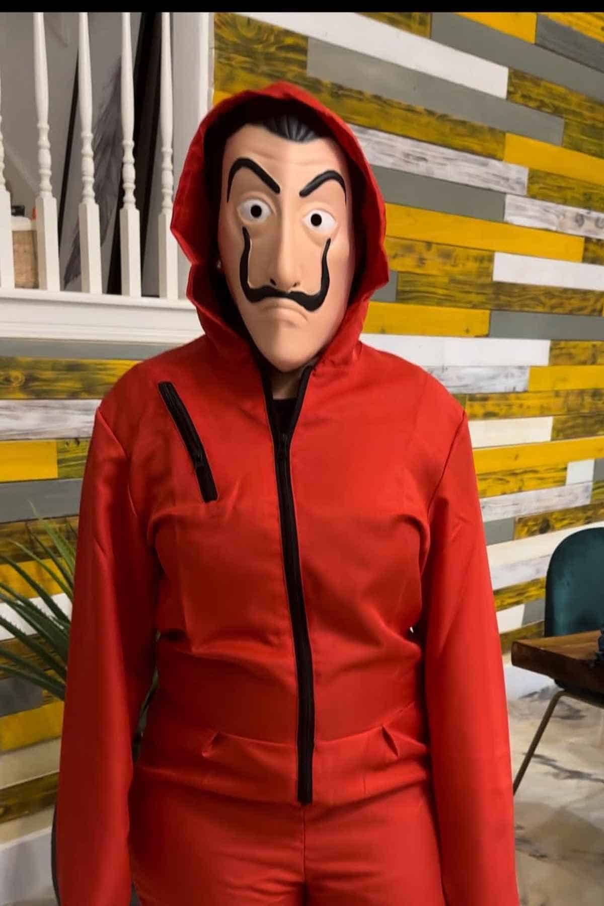 Person wearing a red hooded jumpsuit and a mask, standing indoors in front of a colorful wooden wall.