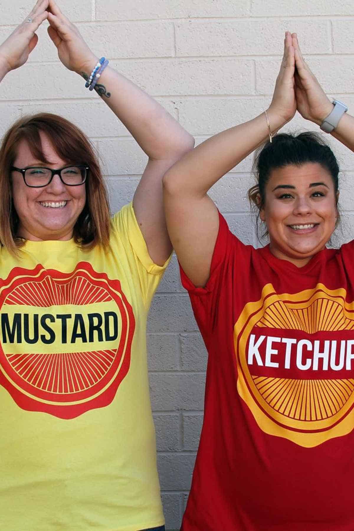 Two women stand side by side in front of a light brick wall, wearing matching shirts labeled "MUSTARD" and "KETCHUP," each raising their arms above their heads.