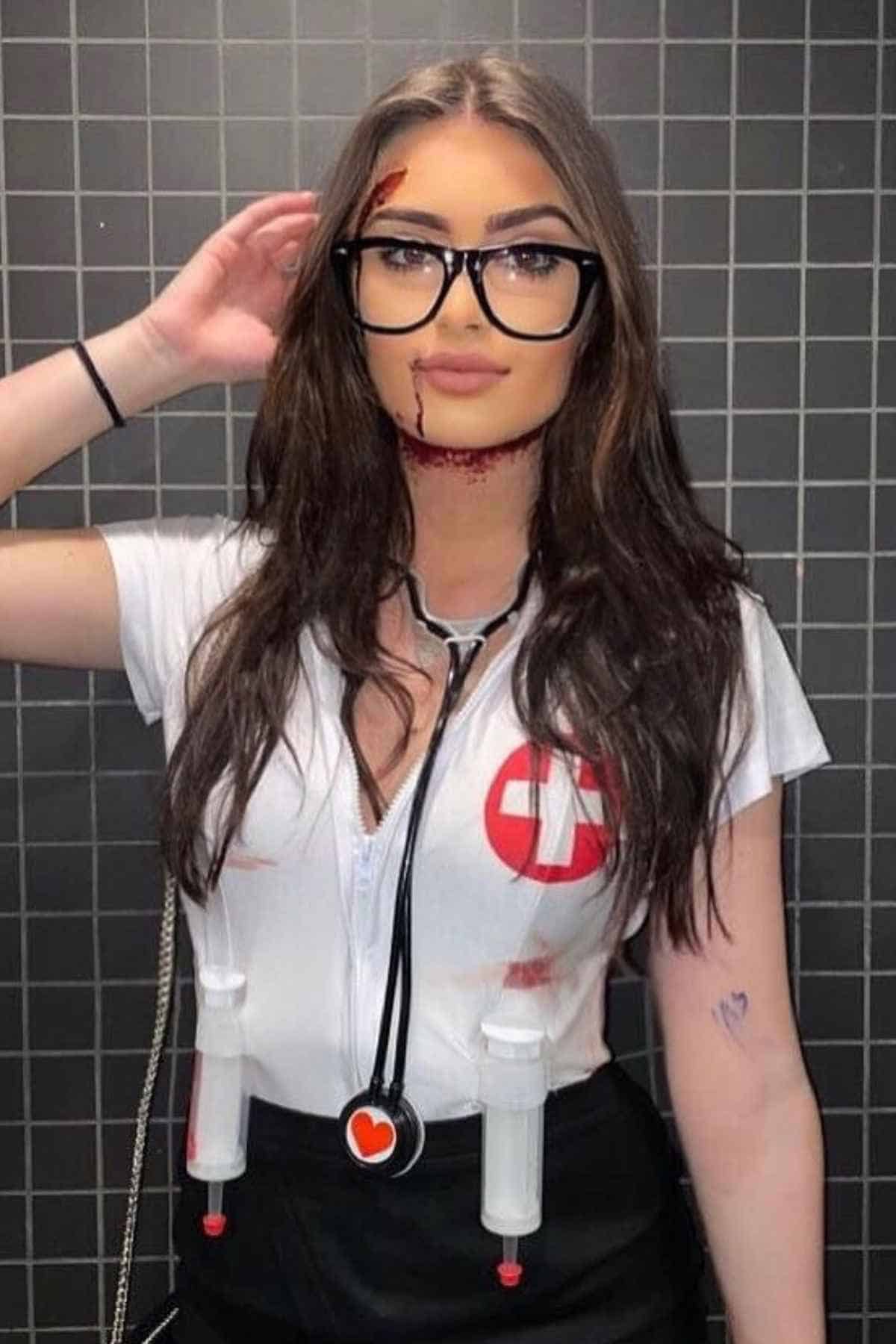 A woman in a nurse costume with fake blood on her face and shirt, wearing glasses and a stethoscope, stands in front of a tiled wall.