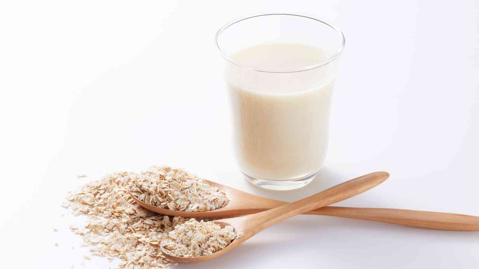 A glass of oat milk next to two wooden spoons filled with dry oats on a white background.
