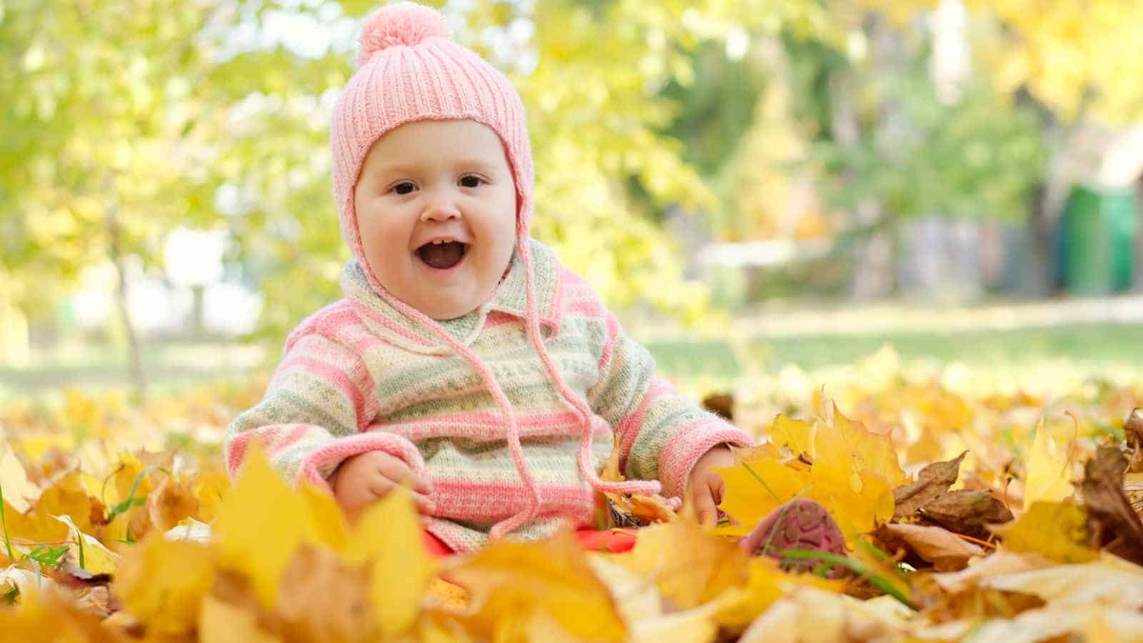 A smiling baby wearing a pink knit hat and striped jacket sits among fallen yellow autumn leaves outdoors.