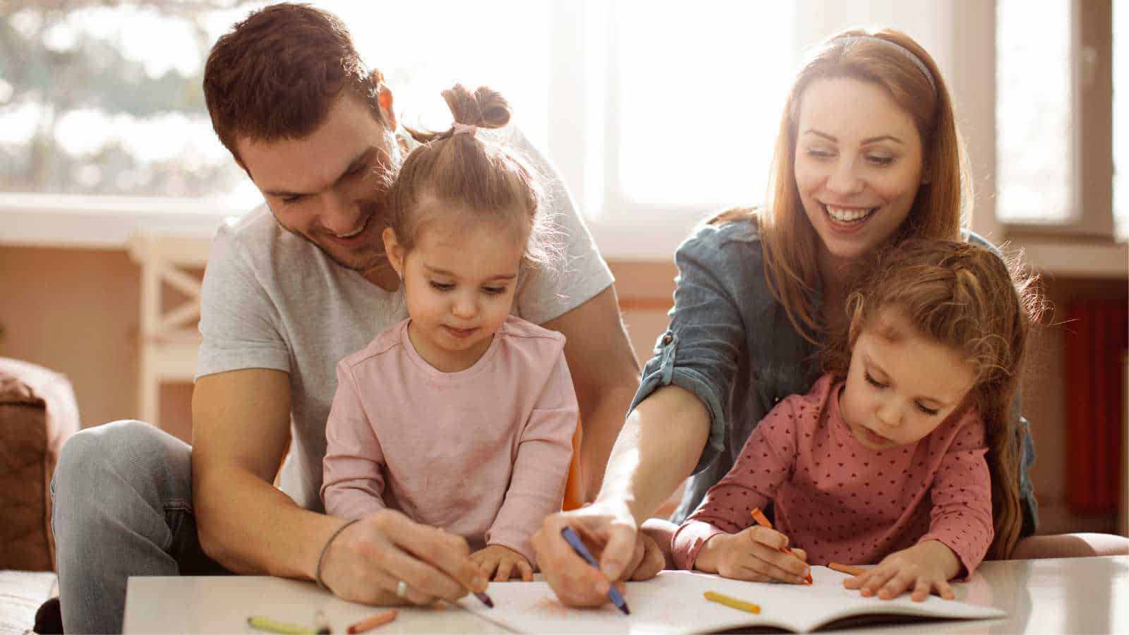 A family of four sits together at a table, with the parents helping their two young daughters color in a coloring book.