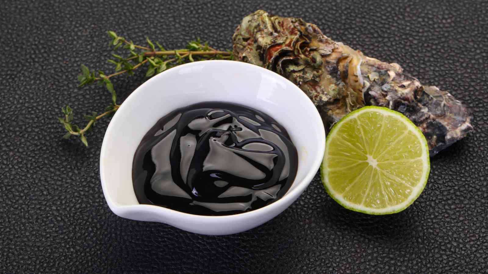 A white bowl of dark oyster sauce is placed beside a halved lime, a sprig of herbs, and an oyster shell on a black textured surface.