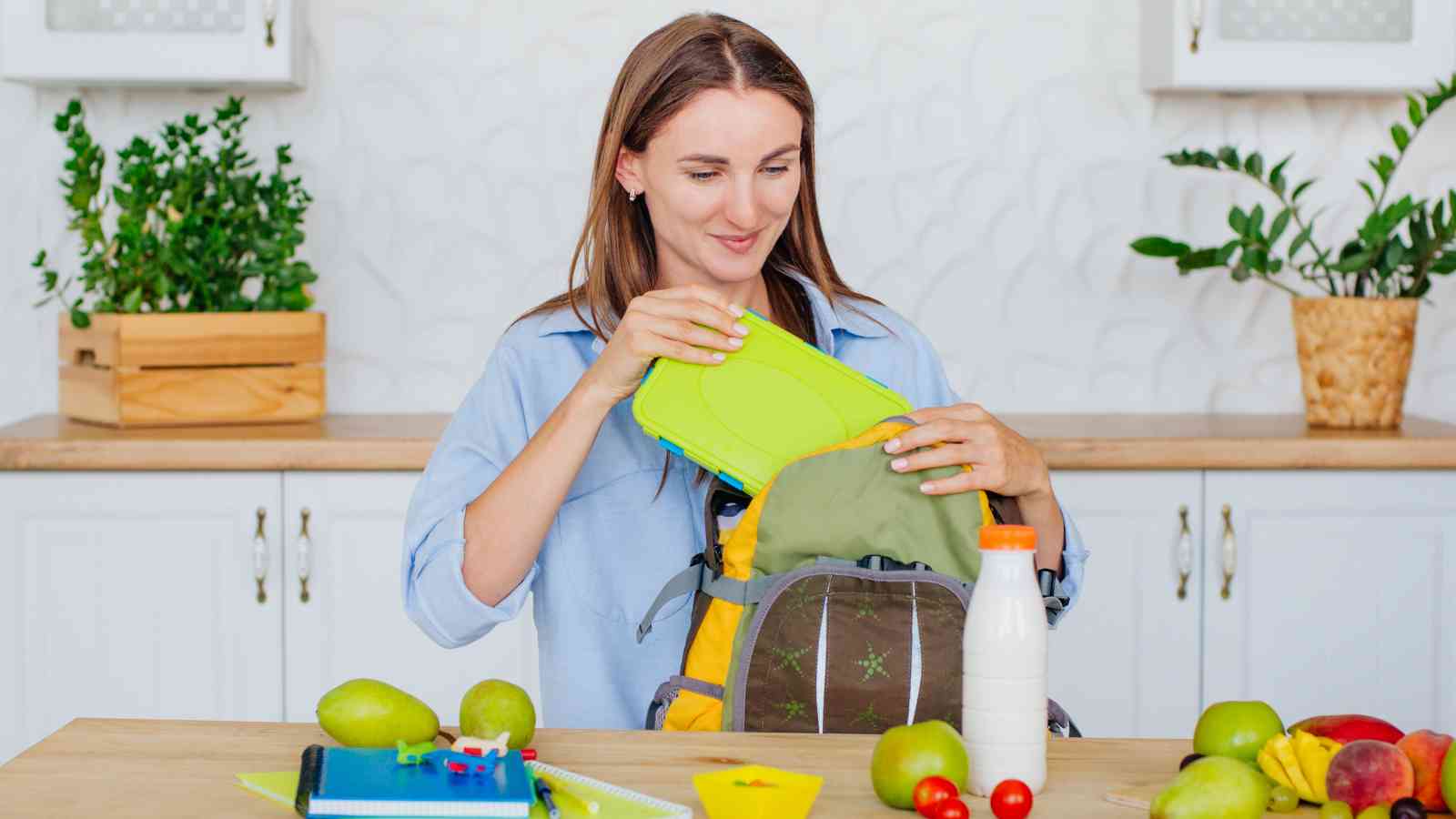 A woman packs a green lunchbox into a backpack on a kitchen counter with fruit, a bottle of milk, and school supplies.