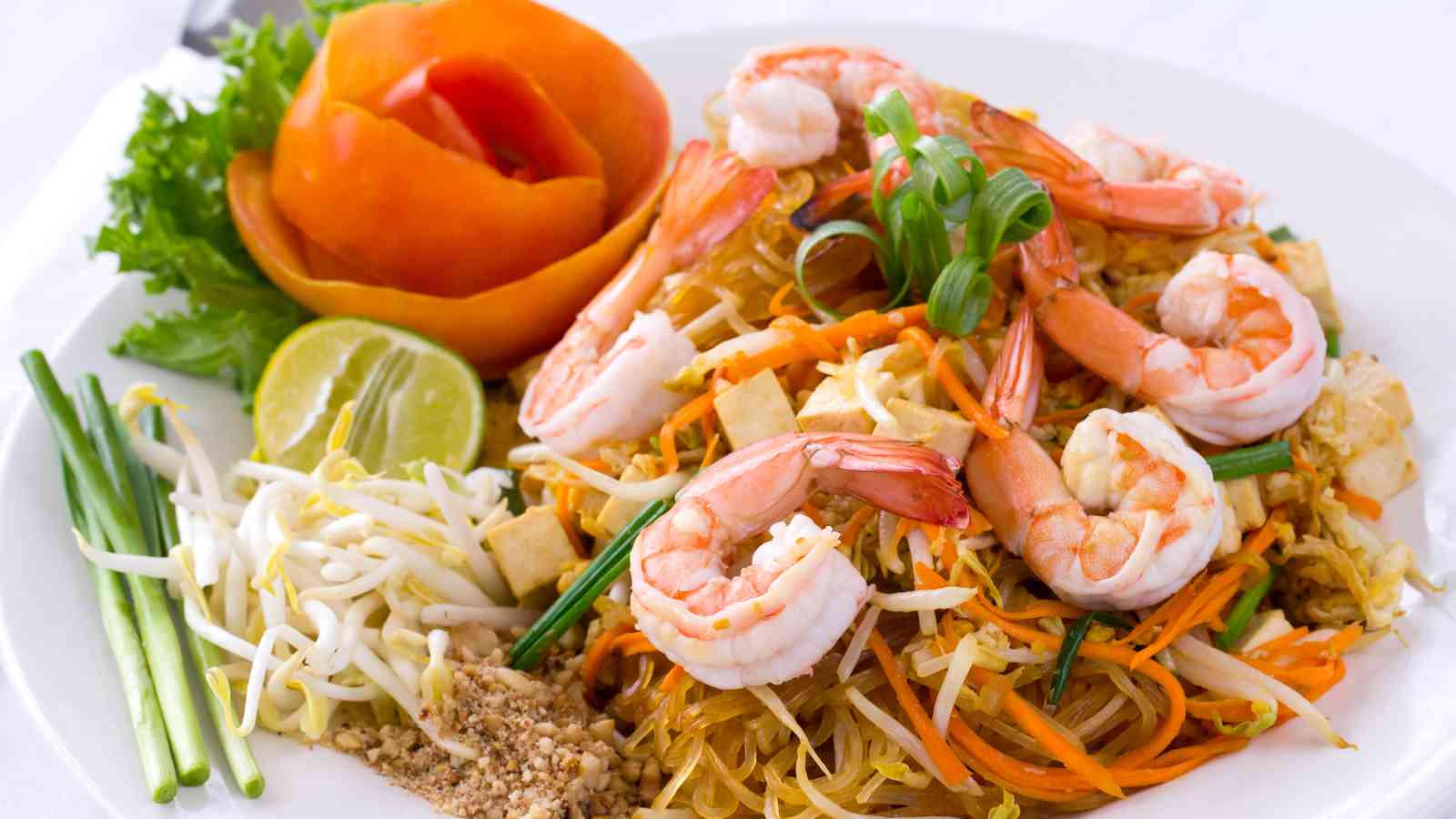 A plate of shrimp pad thai garnished with green onions, lime, bean sprouts, peanuts, and a carved tomato shaped like a rose.