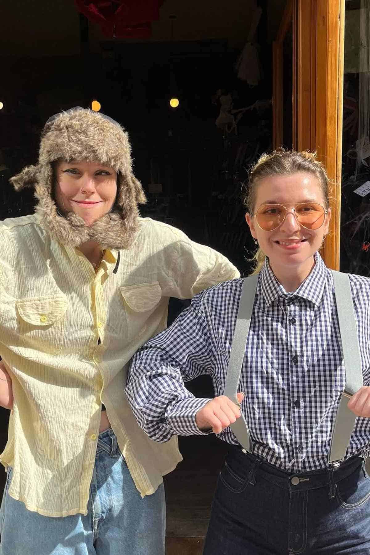 Two people stand side by side in sunlight; one wears a furry hat and yellow shirt, the other wears sunglasses, a checkered shirt, and suspenders.