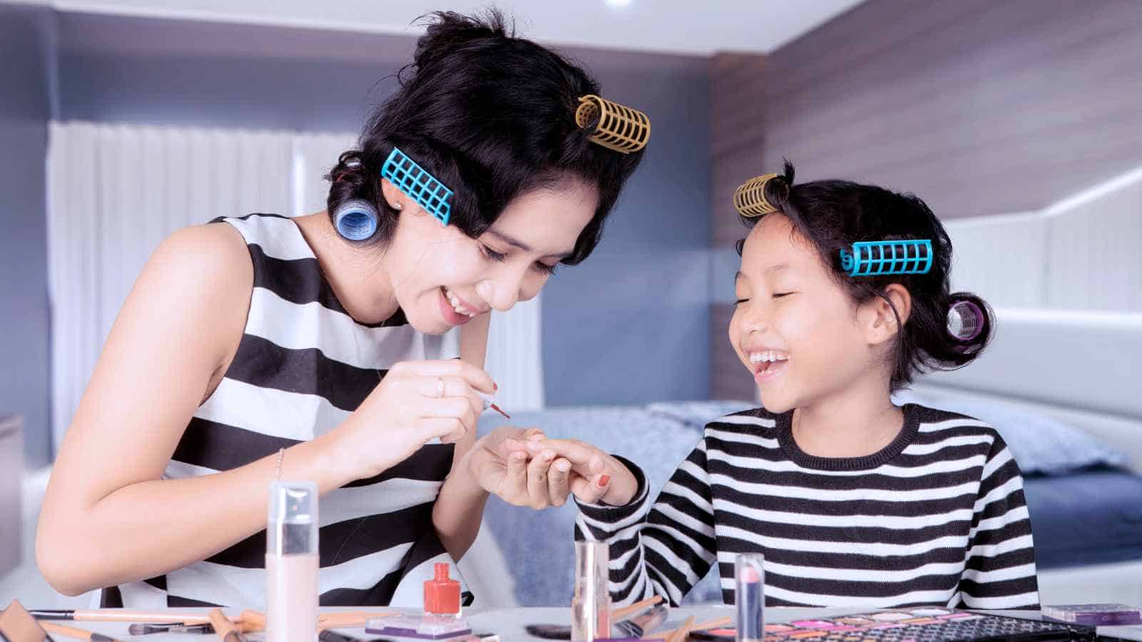 An adult and a child with hair rollers wear matching striped shirts; the adult paints the child’s nails as they sit at a table with makeup items, both smiling.