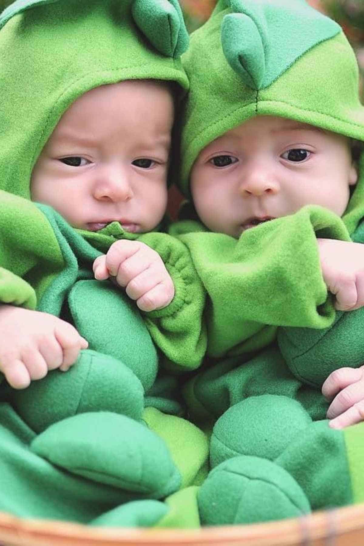Two babies dressed in green pea pod costumes sit close together, facing the camera with neutral expressions.
