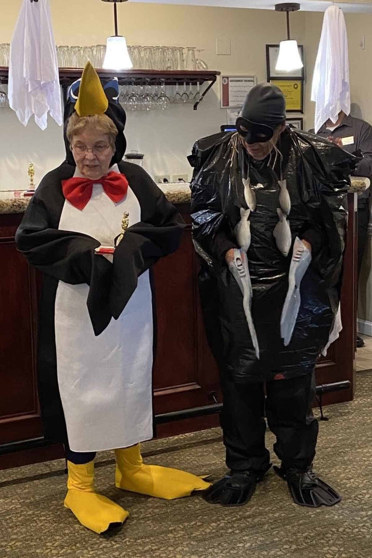 Two people stand indoors in costume; one dressed as a penguin, the other as a black fish or sea creature, both holding white props and standing in front of a bar counter.