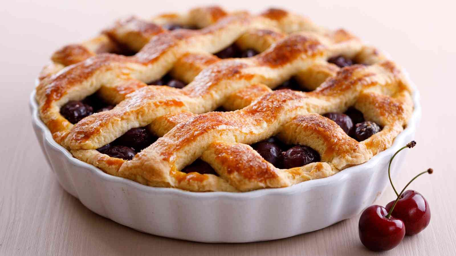 A freshly baked cherry pie with a golden lattice crust sits in a white pie dish, with two cherries placed beside it.