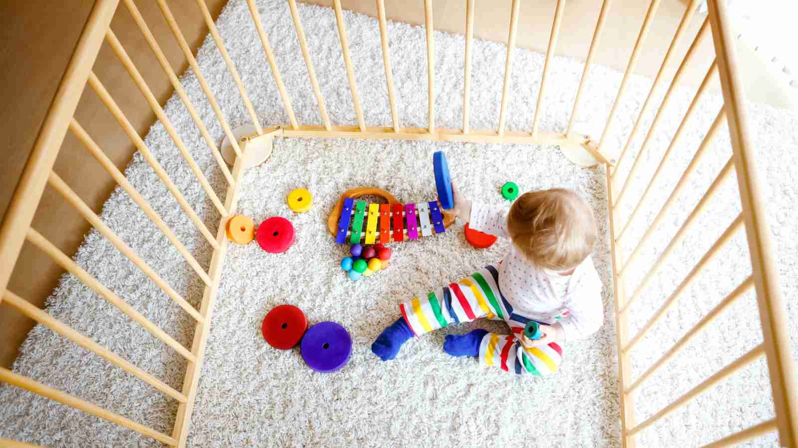 A baby in colorful clothes sits in a playpen on a white carpet, playing with a xylophone and colorful stacking rings.