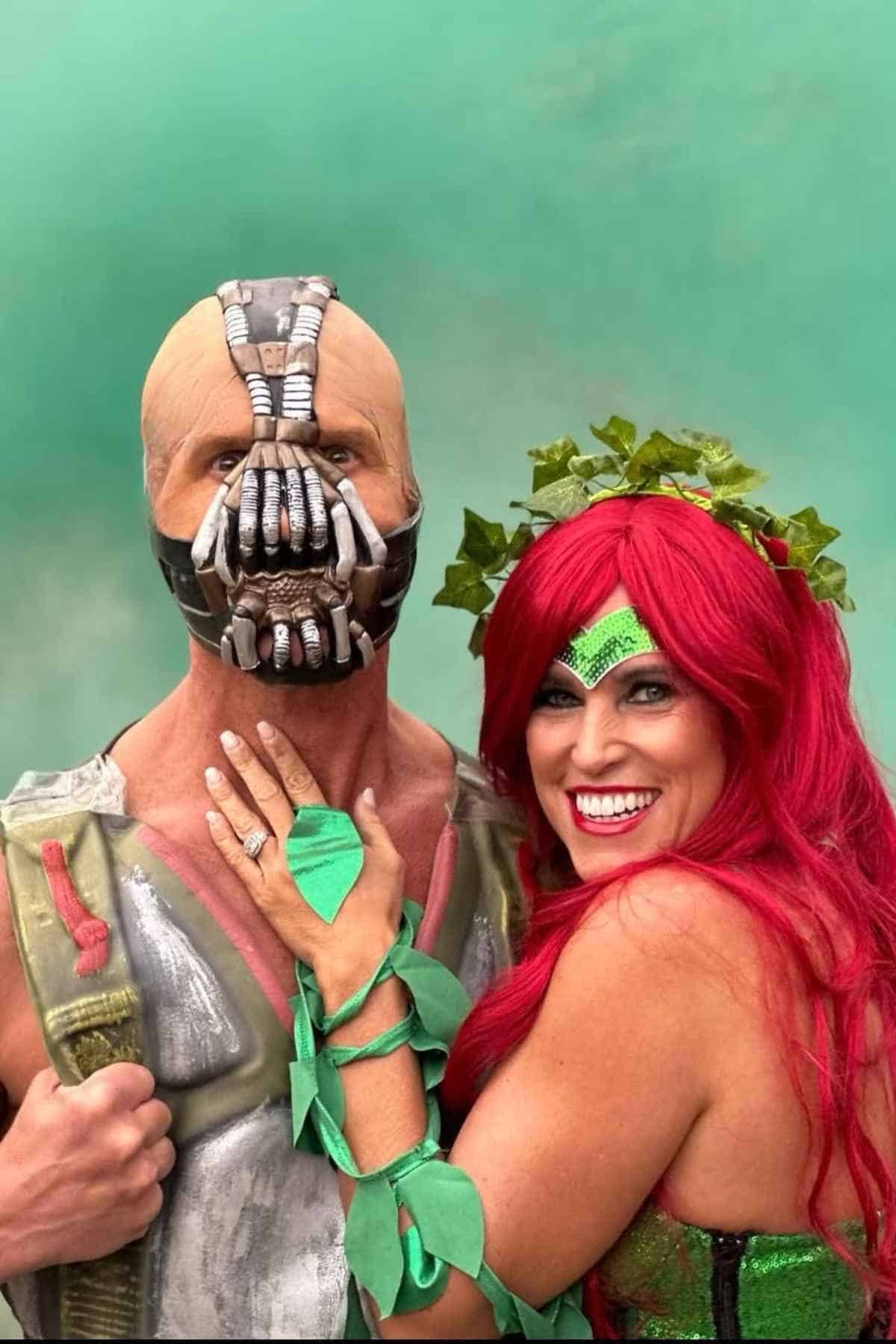 Two people dressed as Bane and Poison Ivy from Batman pose together in costume against a green background.