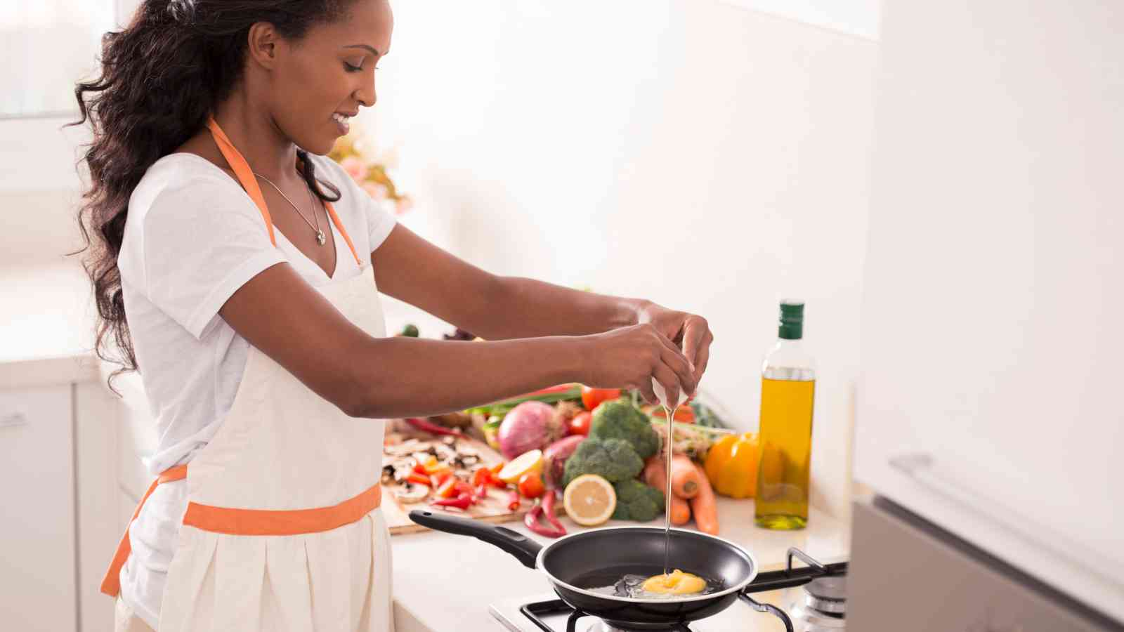 A woman in a white apron cracks an egg into a frying pan on a stove, with various fresh vegetables and a bottle of oil on the counter nearby.