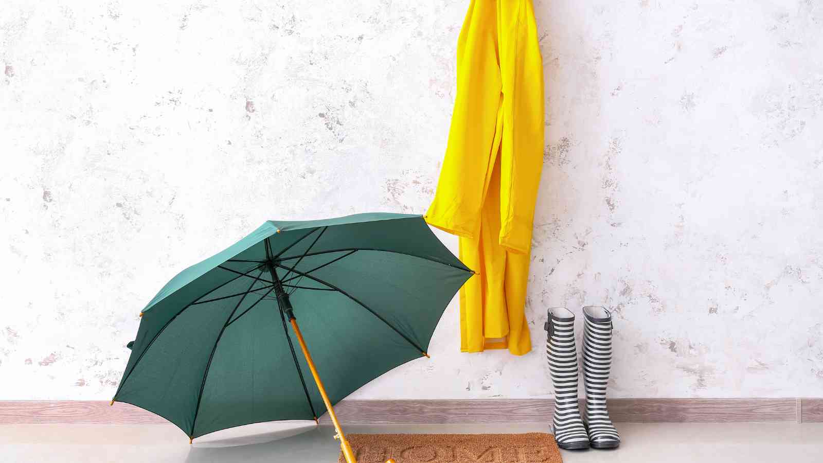 A green umbrella, yellow raincoat, and black-and-white striped rain boots are arranged near a wall with a "home" doormat.