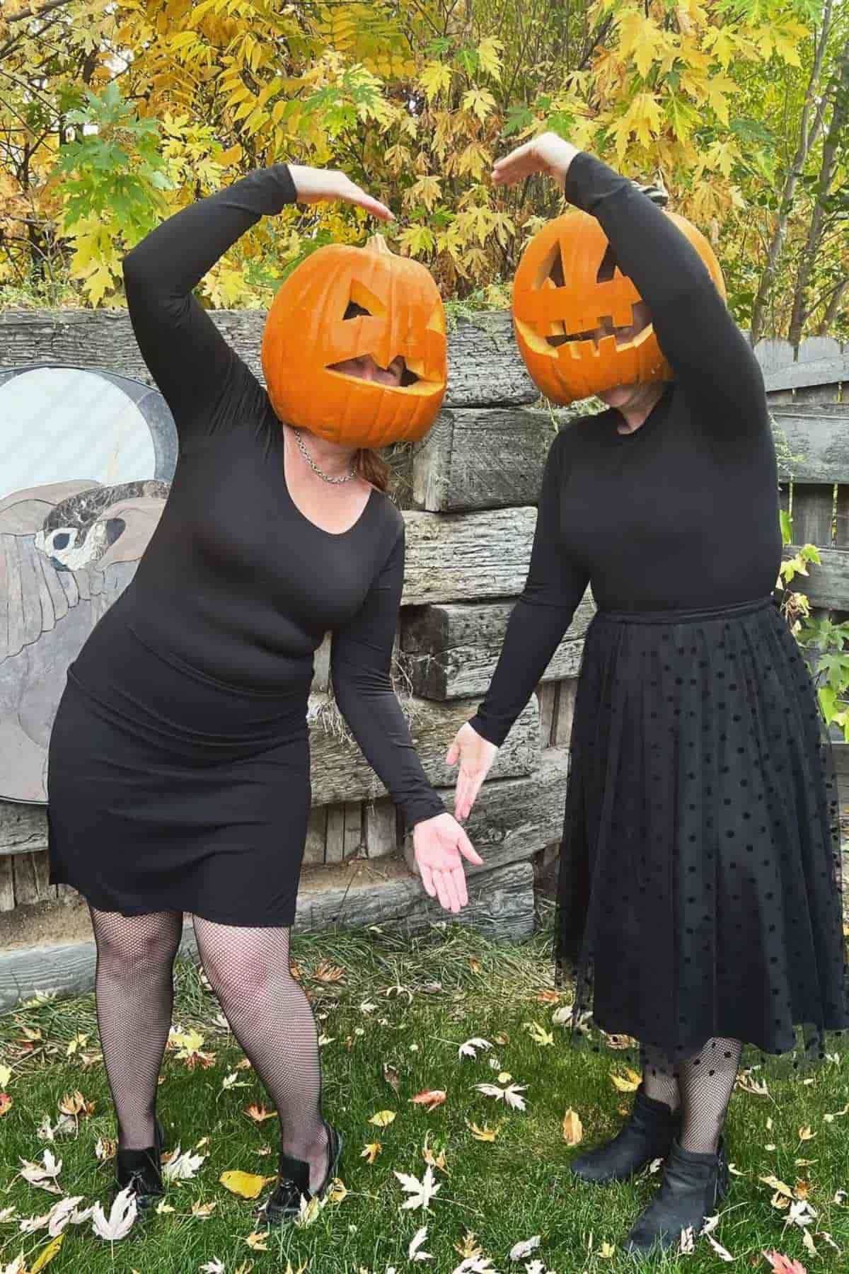Two people wearing black outfits and pumpkin masks pose outdoors, forming a heart shape with their arms. Autumn leaves and a wooden fence are in the background.