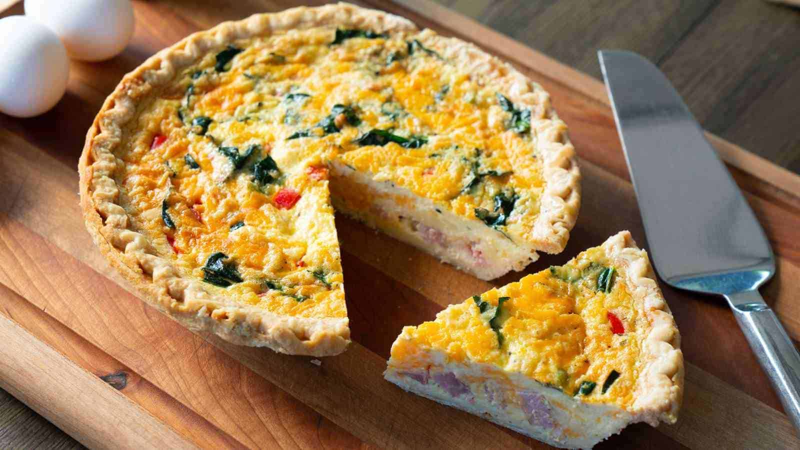 A quiche with spinach and cheese on a wooden board, one slice cut and placed nearby, with a knife and two eggs in the background.