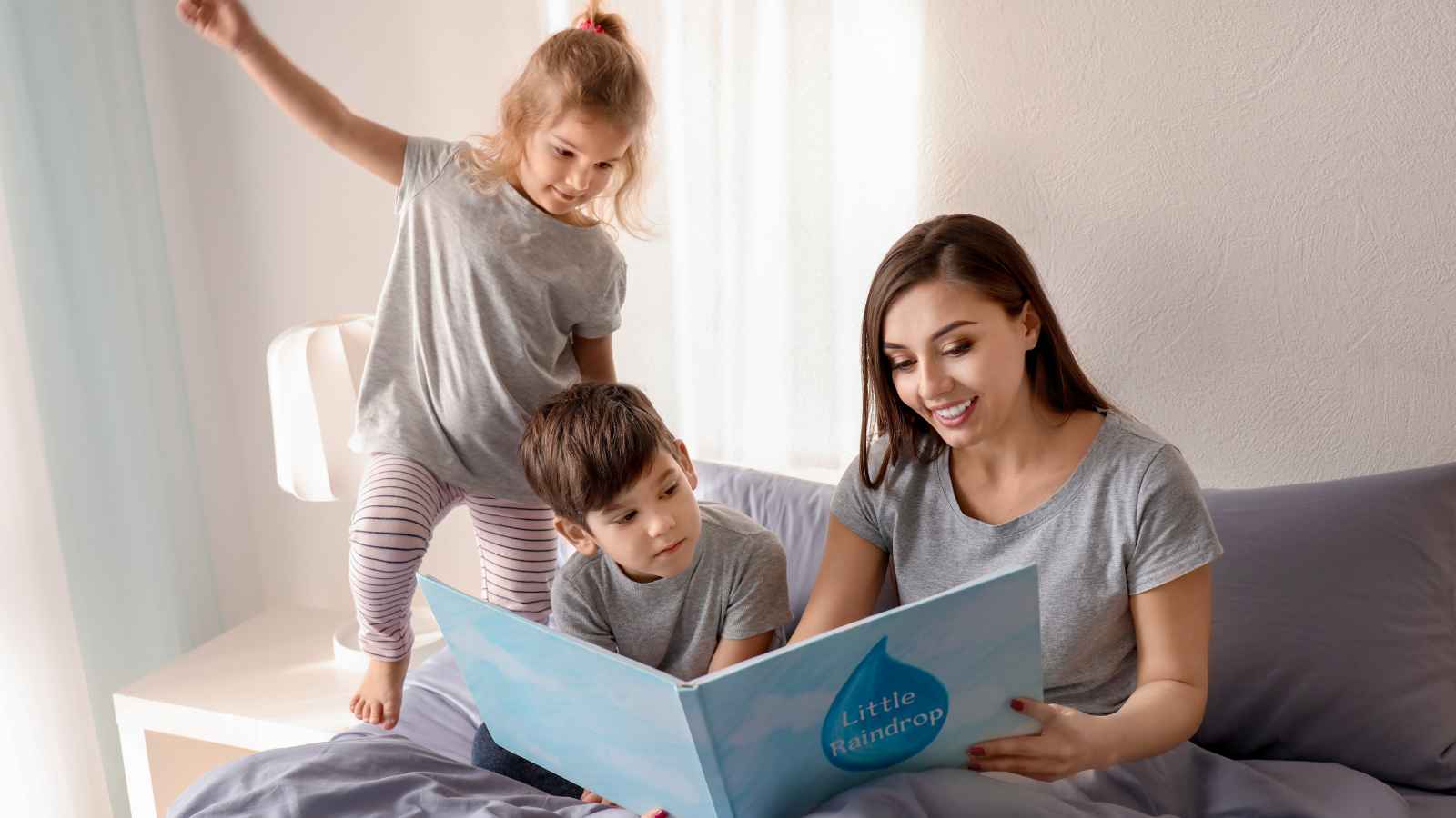 A woman sits on a bed reading a children's book to a young boy while a young girl stands beside them, all wearing gray shirts.
