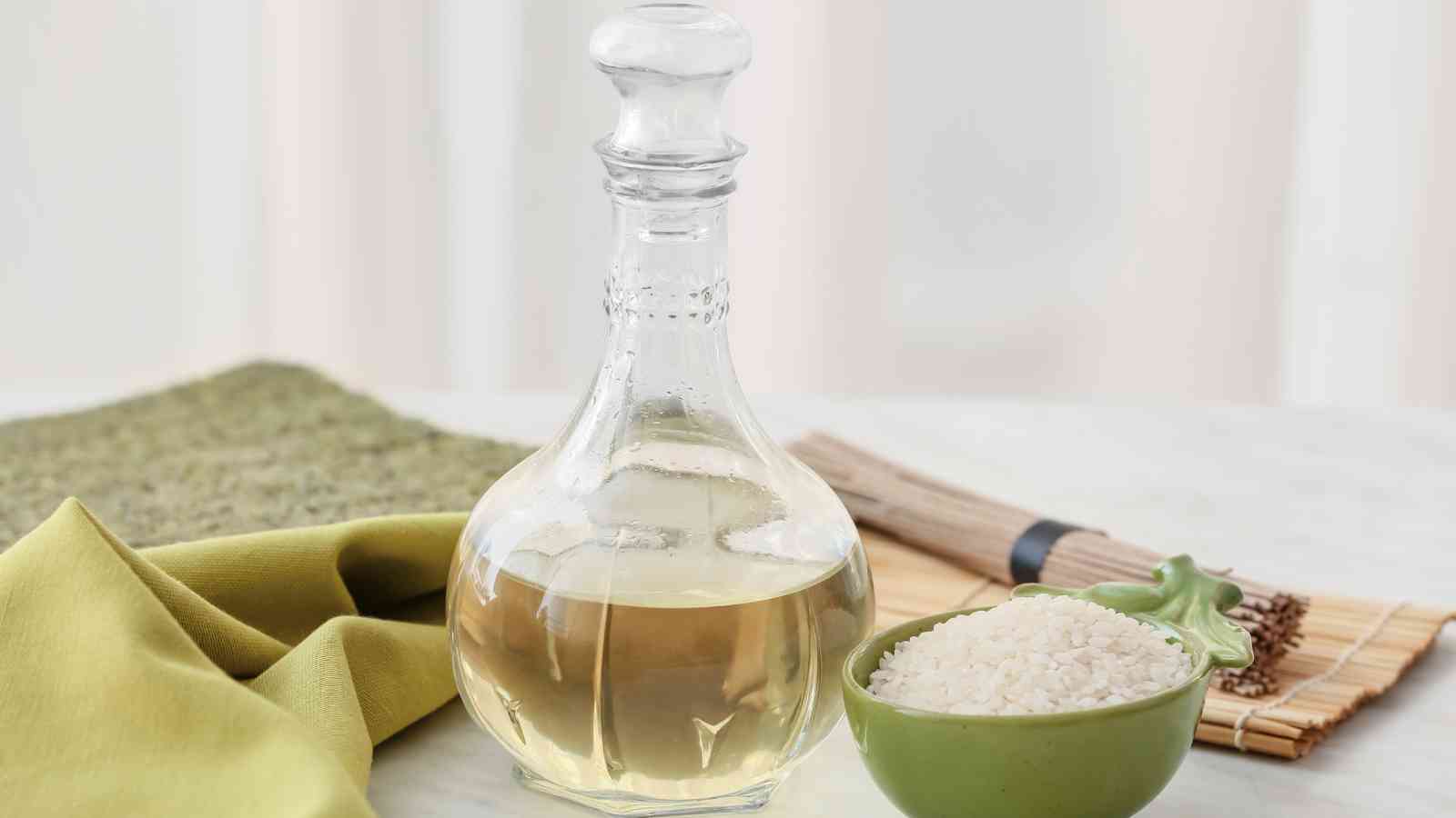 A glass bottle of rice vinegar next to a green bowl of uncooked white rice, a green cloth, and bamboo sushi mat on a white surface.