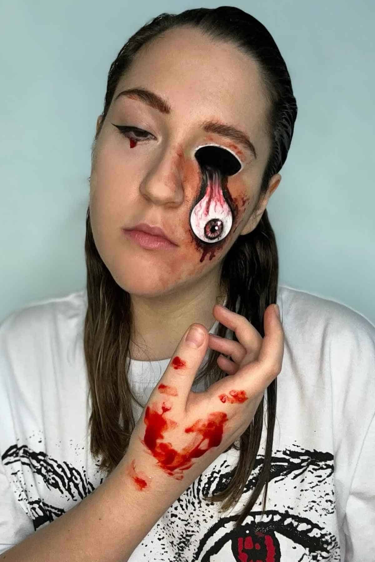 Person with long hair wears makeup resembling a bloodied, melting eye on their cheek and fake blood on their hand, against a plain light background.