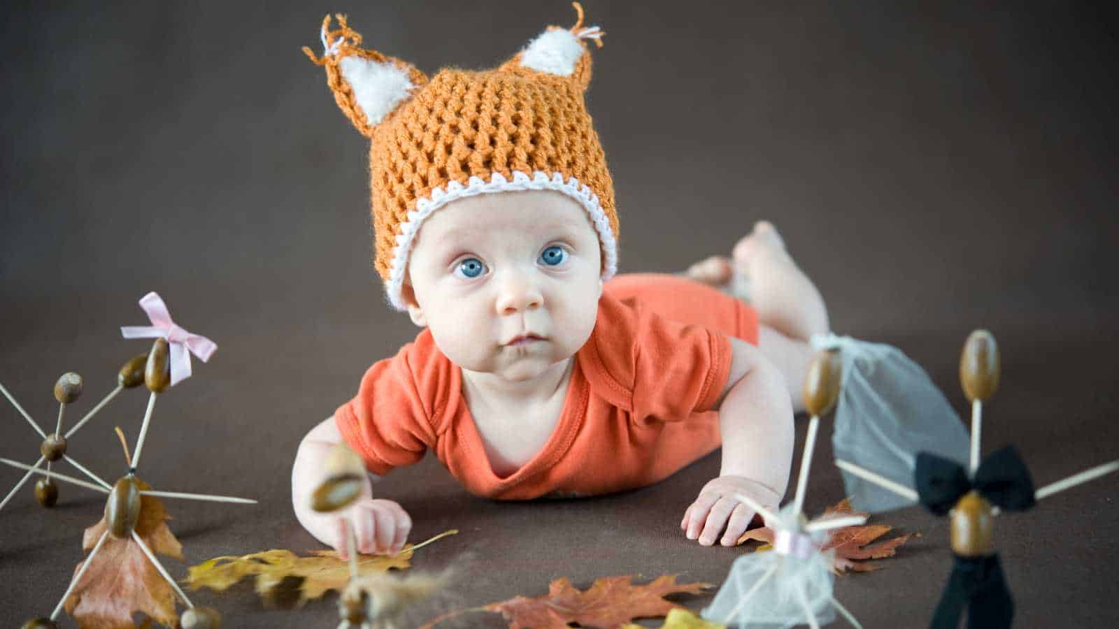 A baby in an orange shirt and a knitted fox hat lies on their stomach, surrounded by autumn leaves and acorn crafts on a brown background.