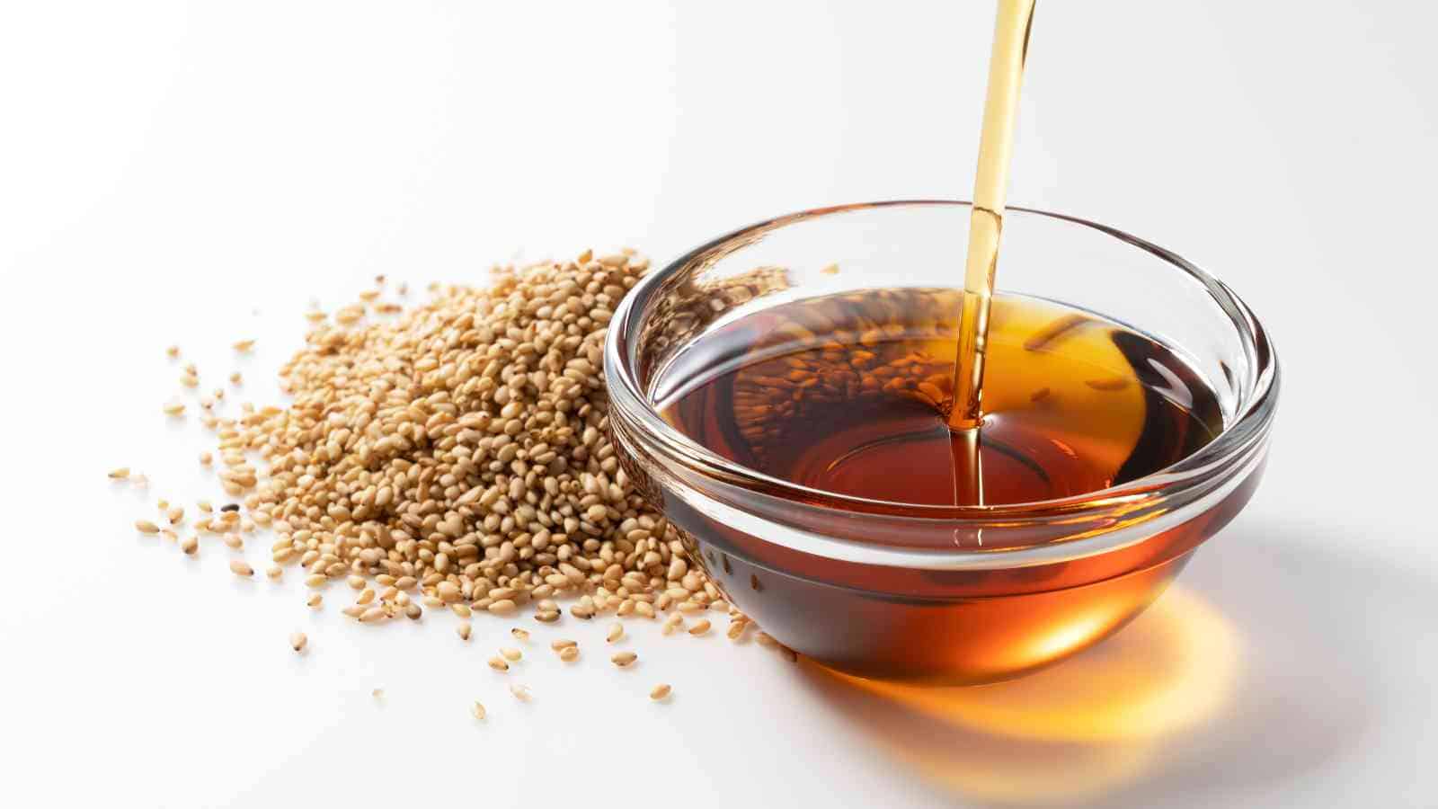 A stream of sesame oil is poured into a glass bowl next to a pile of raw sesame seeds on a white surface.