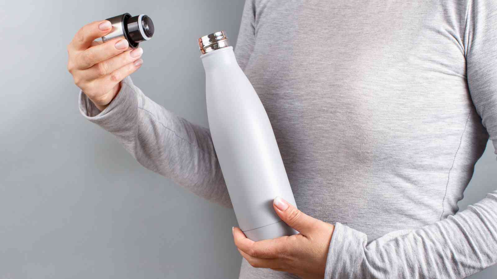 A person in a grey long-sleeve shirt holds a stainless steel water bottle in one hand and removes the black cap with the other hand.