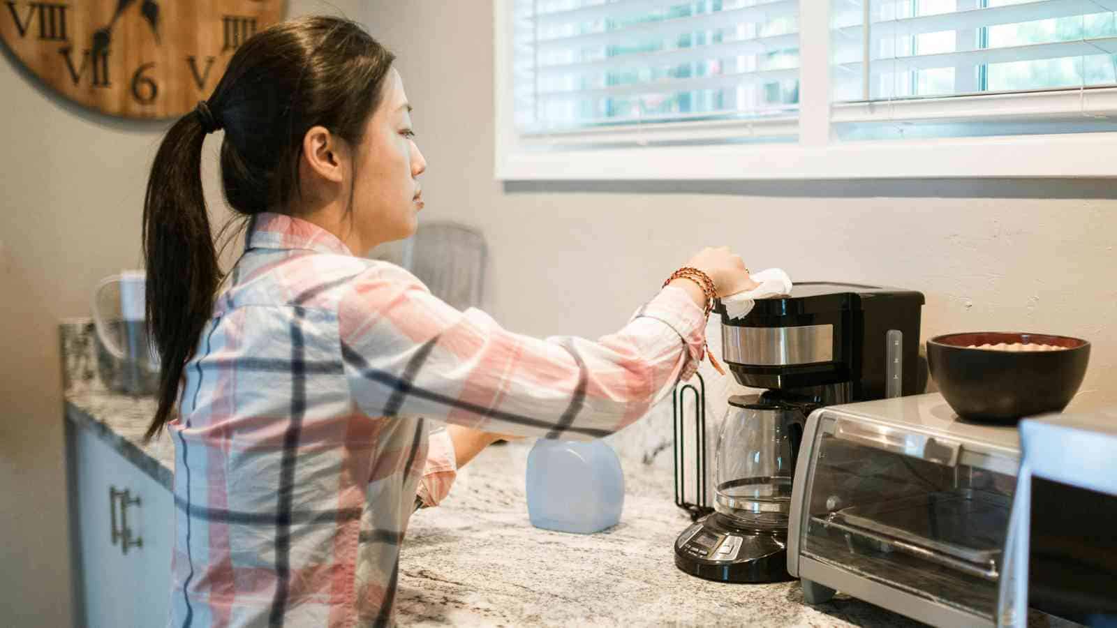 A woman in a plaid shirt prepares coffee using a drip coffee maker on a kitchen counter next to a toaster oven and a bowl.